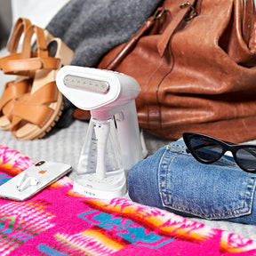 Fridja handheld clothes steamer in white rested on colourful blanket beside Iphone and Airpods and folded jeans