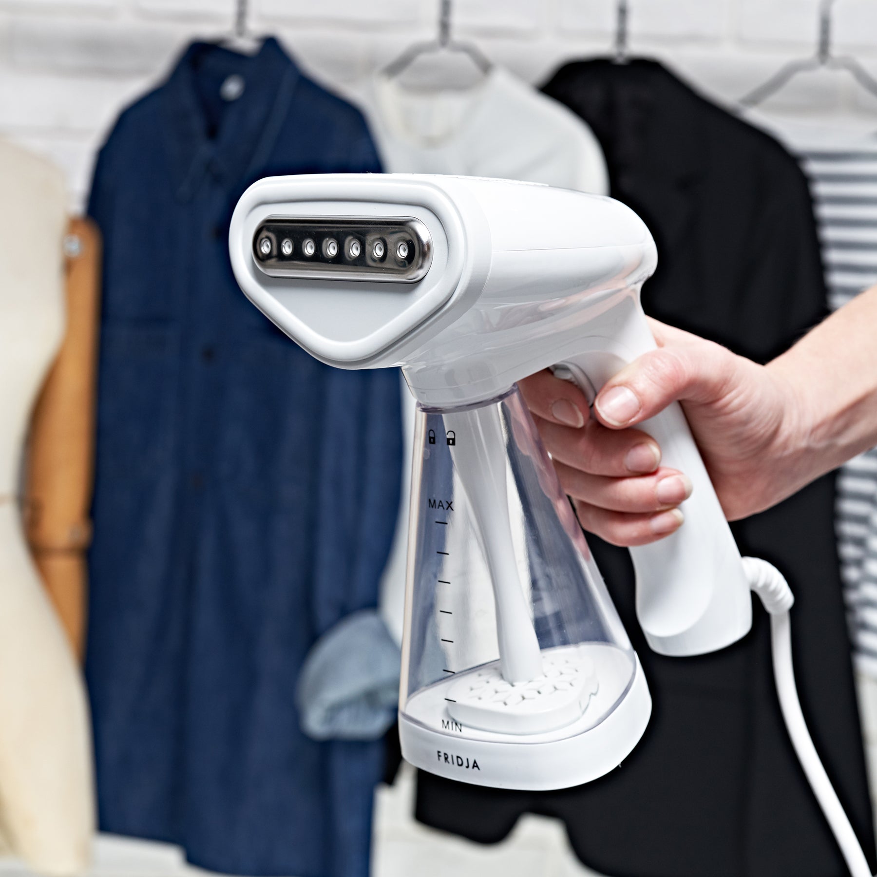 Person holding white handheld clothes steamer from Total Wardrobe Care with hanging clothes out of focus to background