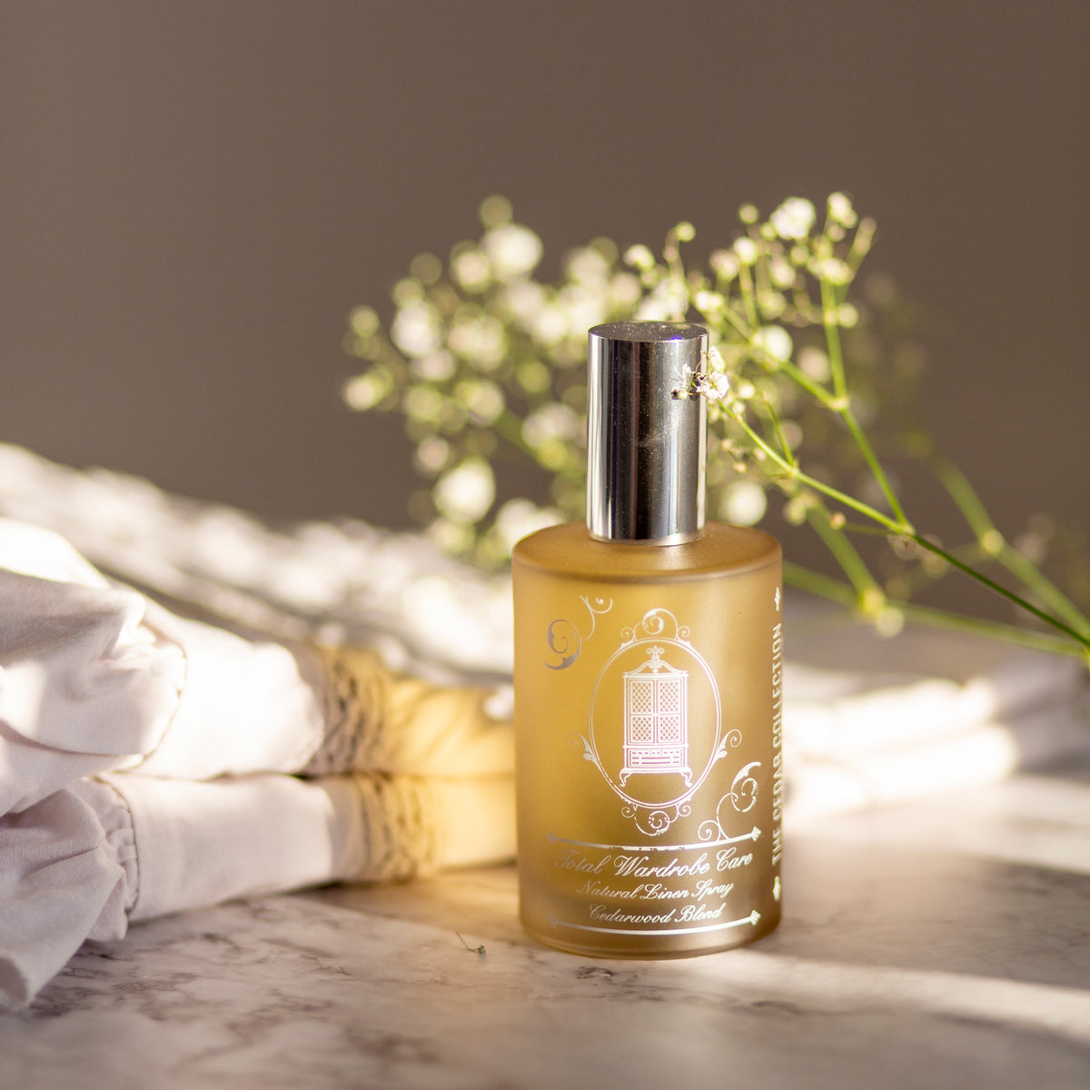 Golden bottle of cedarwood linen spray beside folded linen and flowers to reverse