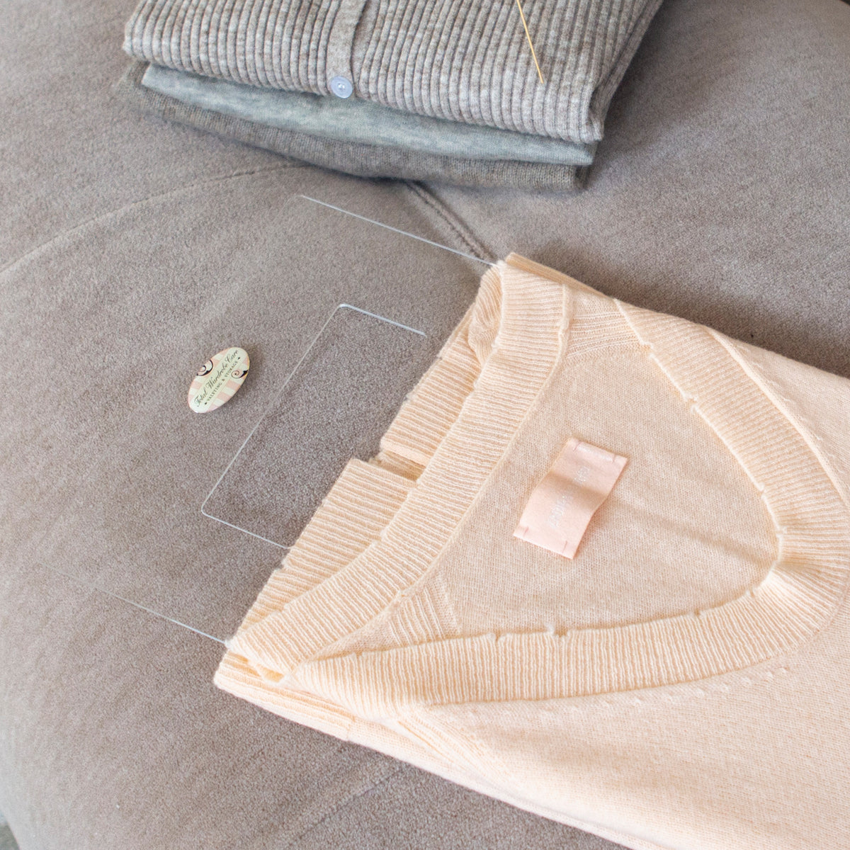Peach cashmere jumper being folded using the Total Wardrobe Care folding palette beside stack of folded clothes